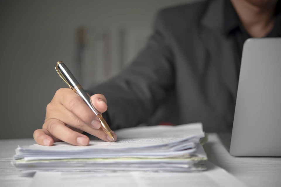 Deux personnes en consultation dans un bureau juridique, l'une en chemise grise tenant un stylo au-dessus d'un dossier ouvert, l'autre en veste beige observant